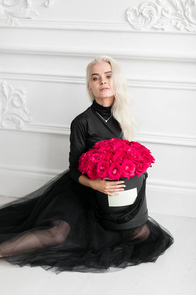 looking at the camera. Young pretty girl with cute face and long blond hair. Woman standing and holding black box with pink roses. Fashion style studio portrait. Luxury interior - Foto, Bild