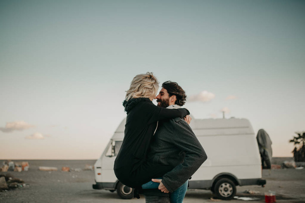 Cool young couple kissing each other outdoors while they are embraced, during a road trip stop, with their van in the background. - Photo, image