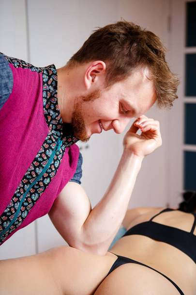Um terapeuta de massagem masculino com barba em um terno vermelho faz uma massagem anti-celulite para uma menina em roupa interior preta com a ajuda de um cotovelo no salão de beleza em uma mesa de massagem.
 - Foto, Imagem