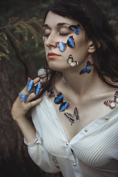 Beautiful art portrait of a naked woman with blue butterflies on her face and hands near the branch of spruce in nature in the forest. Mysterious magical romantic view of girl near the tree branches. - Foto, Imagen