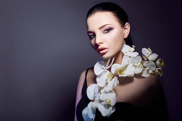 Brunette meisje met orchidee bloemen op het gezicht en de borst, de schoonheid portret van een perfecte make-up, mooie ogen en dikke lippen, jensina op een donkere paarse achtergrond. Gezichts- en huidverzorging. - Foto, afbeelding