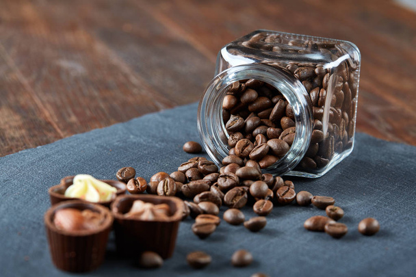Side view of overturned glass jar with coffee beans and chocolate candies on wooden background, selective focus - Φωτογραφία, εικόνα