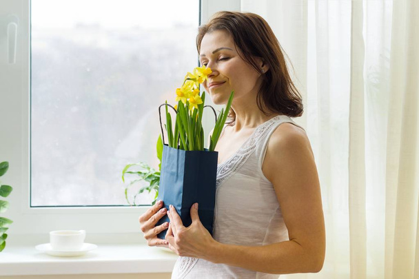 Middle aged woman enjoys a bouquet of yellow spring flowers. At home, near the window - Foto, Imagem