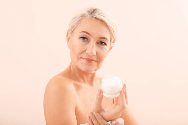 Portrait of beautiful mature woman with jar of cream on light background. Skin care concept - Fotoğraf, Görsel