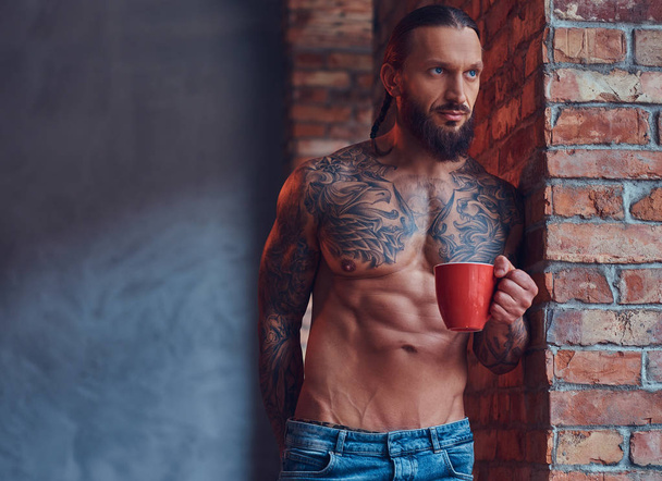 Retrato de un hombre sin camisa tatuado con un corte de pelo elegante y barba, bebe café de la mañana, apoyado contra una pared de ladrillo en una habitación con un interior loft
. - Foto, imagen