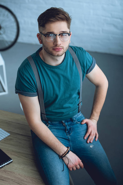 high angle view of handsome young man in eyeglasses sitting on table and looking at camera - Φωτογραφία, εικόνα