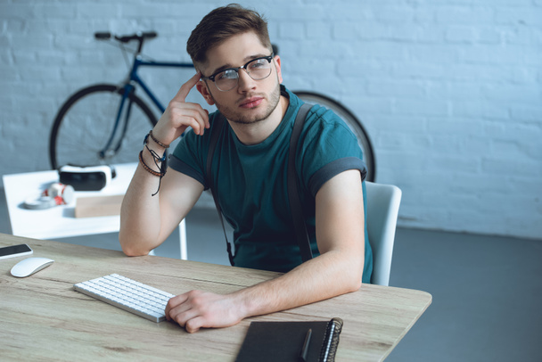 bonito pensativo freelancer em óculos olhando para longe enquanto sentado no local de trabalho
 - Foto, Imagem