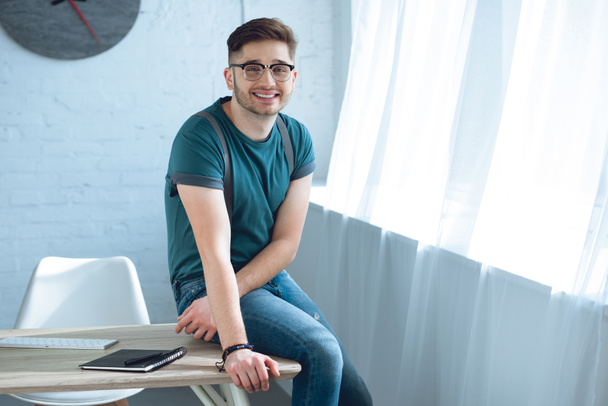 handsome young man in eyeglasses smiling at camera while sitting on table at home office - Photo, image