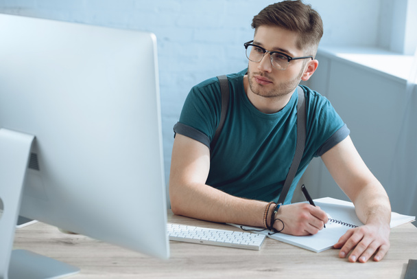 handsome young freelancer in eyeglasses taking notes in notebook while working with desktop computer - Photo, Image