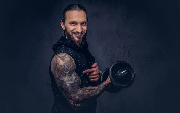 Portrait of a muscular bearded tattoed male with a stylish haircut wearing black sportswear, doing exercise with a dumbbell, isolated on a dark background. - Photo, Image
