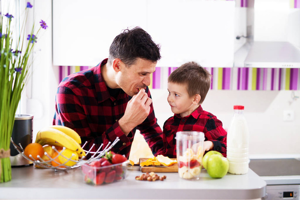 молодой веселый отец и сын в красных рубашках готовят здоровый завтрак с блендером и дегустацией фруктов
  - Фото, изображение