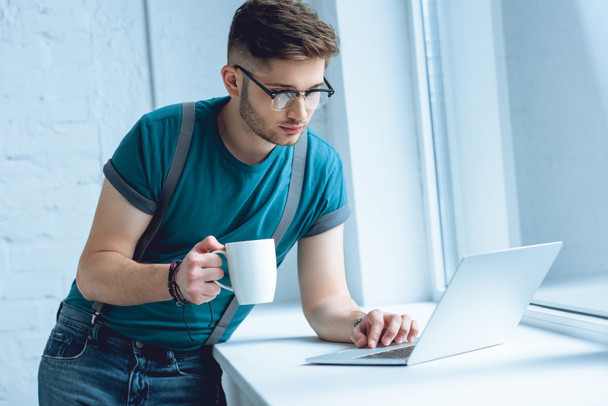 young man in eyeglasses holding cup of coffee and using laptop on windowsill - Foto, Imagen