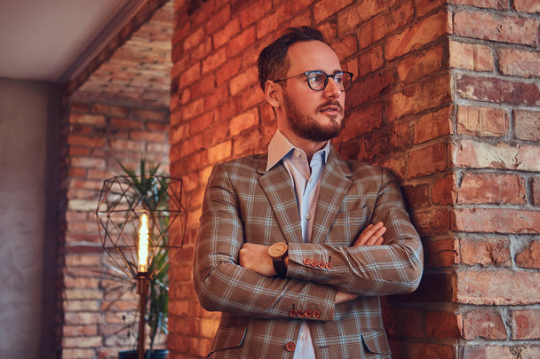 Stylish man in a flannel suit and glasses leaning against a brick wall in a room with loft interior. - Photo, Image