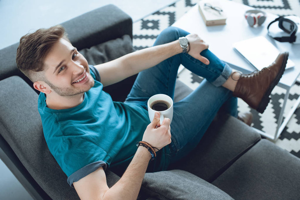 high angle view of handsome young man drinking coffee and smiling at camera at home - Zdjęcie, obraz