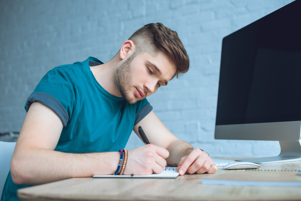 konzentrierter junger Mann schreibt in Notizbuch, während er zu Hause im Büro arbeitet - Foto, Bild
