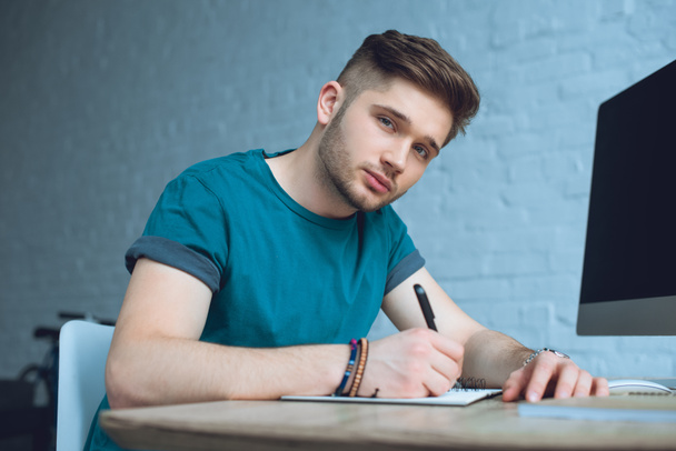 handsome young man taking notes and looking at camera while working at home - Фото, зображення