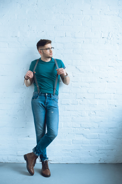 Handsome young man wearing wearing jeans with suspenders and standing by white wall - Fotó, kép