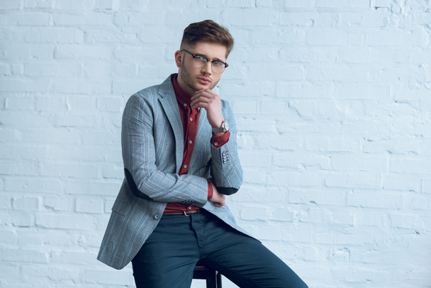 Hombre joven guapo con traje y gafas sentado frente a la pared de ladrillo
 - Foto, Imagen
