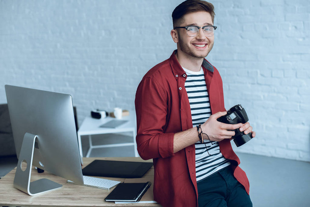 Happy freelancer holding camera by working table with computer - Foto, afbeelding