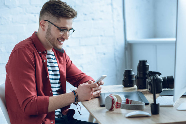 Счастливый фрилансер с помощью смартфона сидит за рабочим столом
 - Фото, изображение