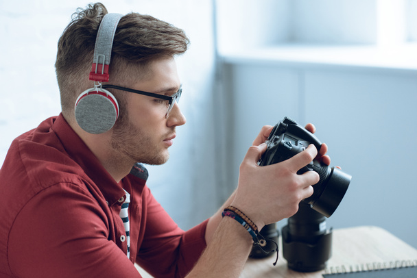 Young bearded man in headphones holding camera by table - Fotografie, Obrázek