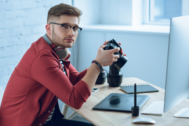 Молодой фотограф держит профессиональную камеру за рабочим столом
 - Фото, изображение