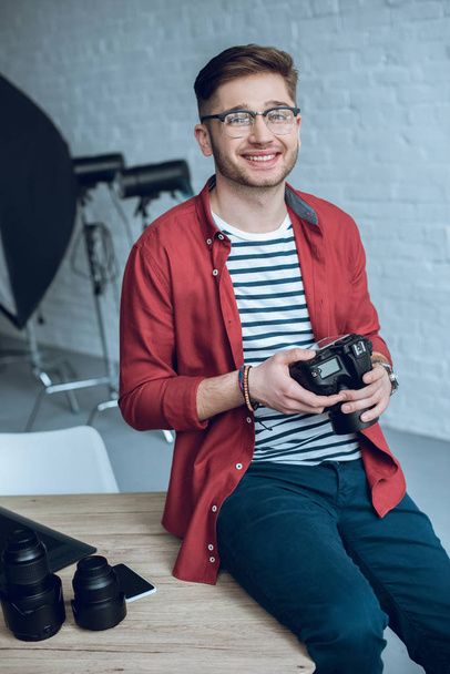 Sonriente hombre sosteniendo la cámara y sentado en la mesa con el ordenador en la oficina en casa
 - Foto, imagen