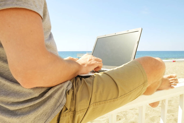 Young man in grey shirt sitting at the beach, working on laptop. Blogger, writer, coder, remote worker surfing browsing researching on notebook. Wireless internet on sea shore. Background, copy space - Photo, Image