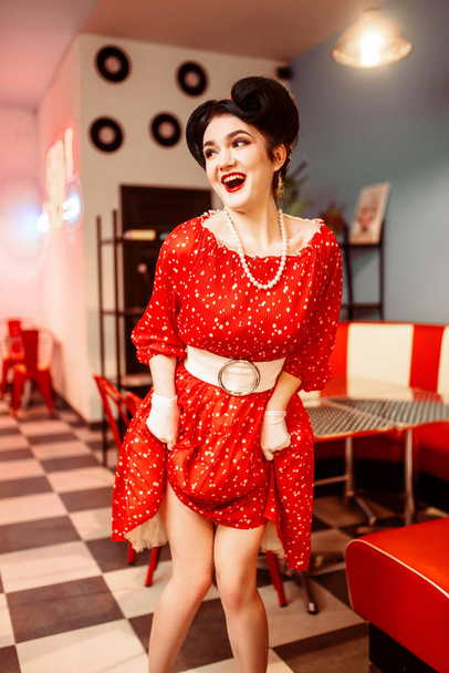 華やかなピンは、レトロなカフェのインテリアでポーズをとって赤いドレスで幸せな女性  - 写真・画像