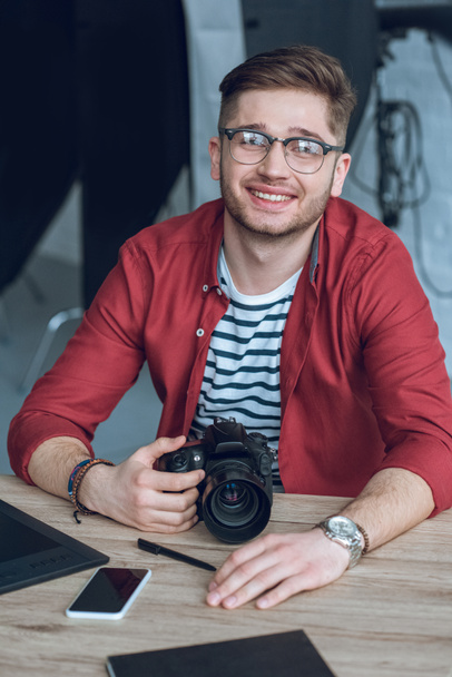 Usmívající se muž nosí brýle a držení fotoaparátu u stolu s počítačem - Fotografie, Obrázek