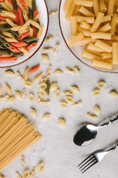 Draufsicht auf verschiedene rohe Pasta und Besteck auf chaotischer Betontischplatte - Foto, Bild