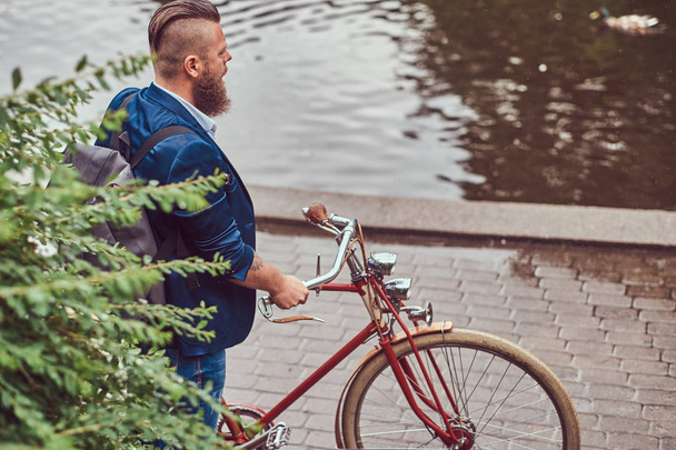 Homme barbu avec une coupe de cheveux élégante habillé en vêtements décontractés avec un sac à dos, debout avec un vélo rétro près de la rivière dans un parc de la ville
. - Photo, image