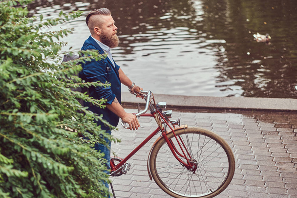 都市公園における川の近くのレトロな自転車の側に立って、バックパックでカジュアルな服に身を包んだスタイリッシュな散髪ひげを生やした男性. - 写真・画像