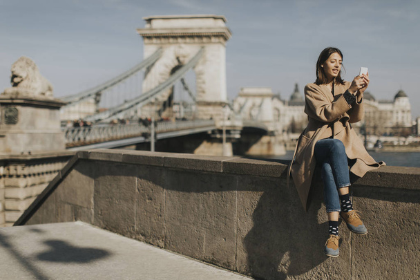 Young woman using mobile phone with Chain bridge at background in Budapest, Hungary - Photo, Image