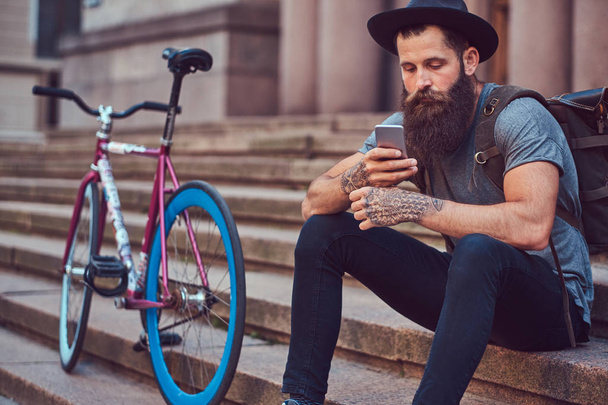 Een knappe hipster reiziger met een stijlvolle baard en tattoo op zijn armen gekleed in casual kleding, zittend op de stappen, met behulp van de telefoon, rust na een fietstocht. - Foto, afbeelding