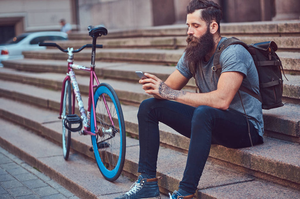 Un guapo viajero hipster con una elegante barba y tatuaje en sus brazos vestido con ropa casual, sentado en las escaleras, usando el teléfono, descansando después de un paseo en bicicleta
. - Foto, imagen