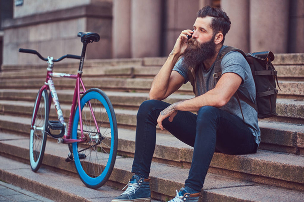 Un beau voyageur hipster avec une barbe élégante et un tatouage sur les bras habillé en vêtements décontractés, assis sur les marches, utilisant le téléphone, se reposant après une balade à vélo
. - Photo, image