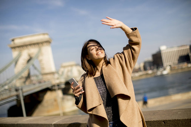 Mujer joven sosteniendo el teléfono móvil con el puente de cadena en el fondo en Budapest, Hungría
 - Foto, Imagen