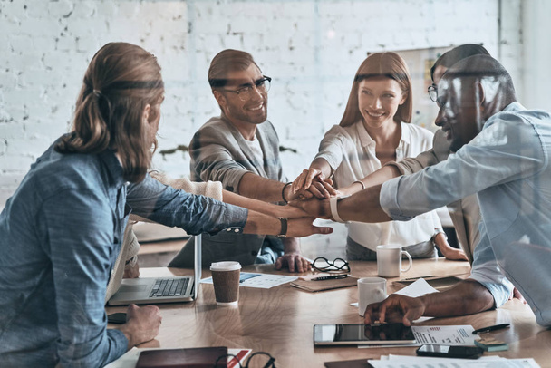 Victory!  Diverse group of business colleagues holding hands on top of one another in a symbol of unity while working behind the glass wall in the board room - 写真・画像