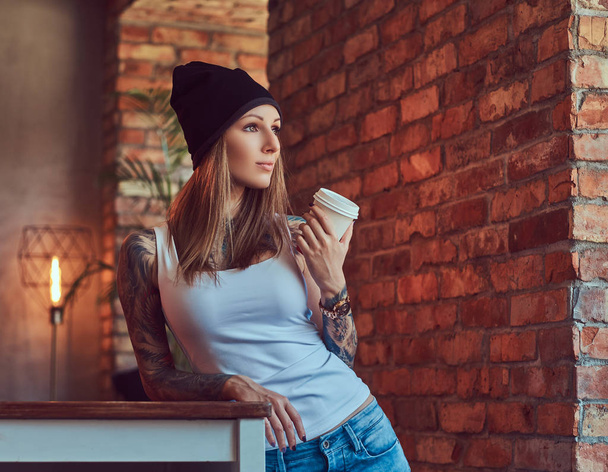 eine tätowierte sexy Blondine in einem T-Shirt und einem Hut mit Tasse Kaffee in einem Raum mit Loft-Interieur. - Foto, Bild