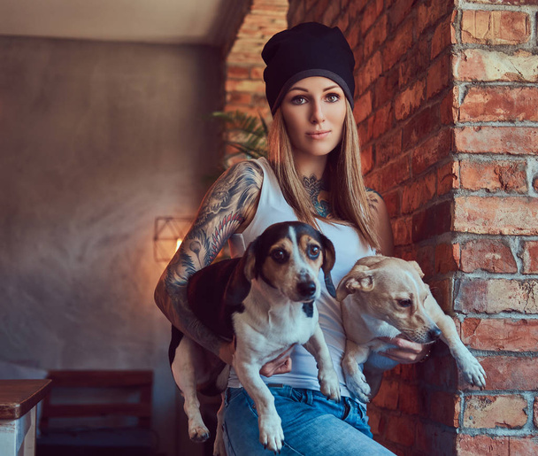 A stylish tattoed blonde female in t-shirt and jeans holds two cute dogs. - Photo, Image