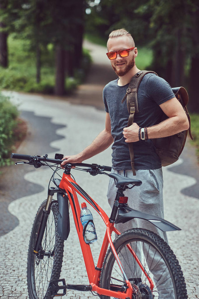 Un hombre pelirrojo guapo con un corte de pelo elegante y barba vestida con ropa deportiva y gafas de sol camina por el parque con una bicicleta y una mochila. - Foto, Imagen
