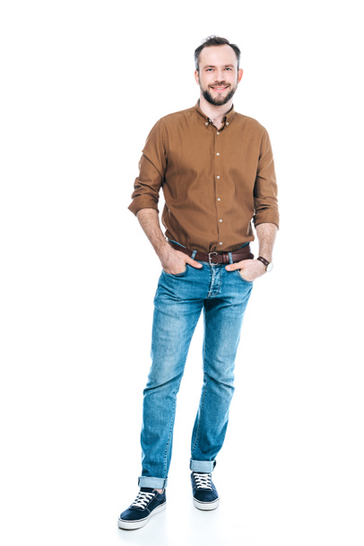 full length view of handsome bearded man standing with hands in pockets and smiling at camera isolated on white - Fotoğraf, Görsel