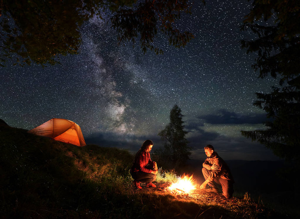 Romantic male and female tourists warming themselves around the fire at night. Camping in the mountains near the trees and illuminated orange tent. - Photo, Image