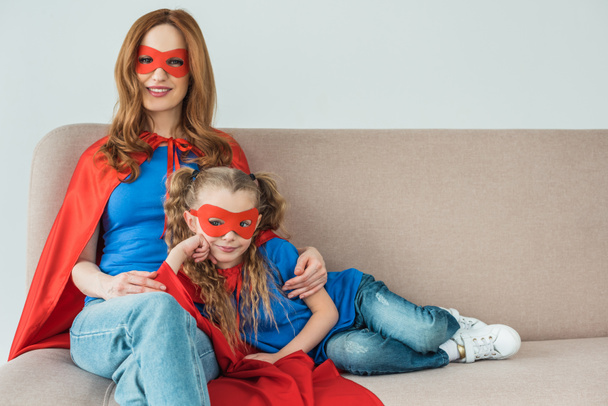 happy mother and daughter in superhero costumes sitting on sofa and smiling at camera - Photo, Image