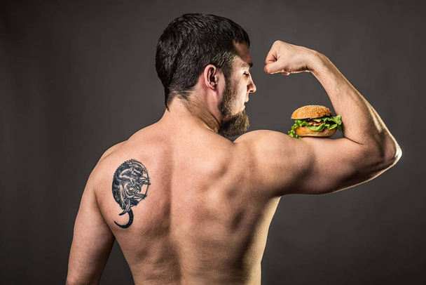 Young man bodybuilder holding burger, view from behind. - Zdjęcie, obraz