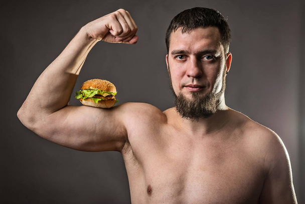 Young man bodybuilder holding burger, unhealthy eating habits. - Valokuva, kuva