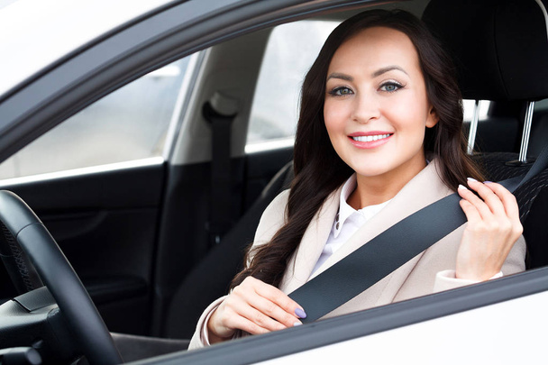 Retrato de joven sonriente, feliz, bonita mujer conductor tirando del cinturón de seguridad dentro de coche blanco
. - Foto, Imagen