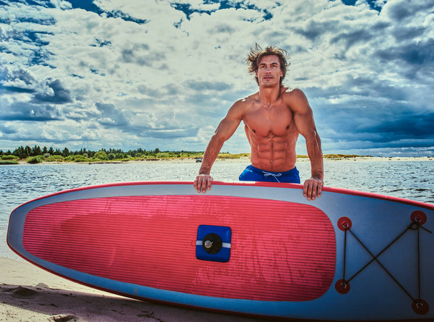 Surffaaja mies, jolla on lihaksikas vartalo ja surffilauta rannalla.
. - Valokuva, kuva