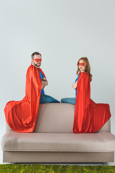 super couple in masks and cloaks sitting on sofa and looking at camera - Zdjęcie, obraz